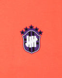 UNDEFEATED SP26 EMBLEM S/S TEE-CORAL